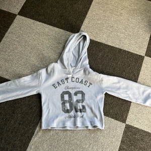 Ljusblå hoodie från Lindex - Säljer en ljusblå hoodie från Lindex med trycket 'East Coast Champions 82 Athletic Dept' på framsidan. Tröjan har en skön huva och långa ärmar, perfekt för en avslappnad stil. Passar bra för ungdomar som gillar sportig look. Helt ny har bara testat