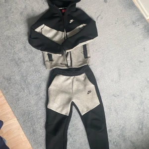 Nike tech fleece set - Säljer en snygg hoodie och matchande byxor från Nike i svart och grått. Hoodien har dragkedja och Nike-logga på bröstet. Byxorna har elastisk midja och logga på benet. Perfekt för en avslappnad stil. Den är äkta, har använts fåtal gånger!