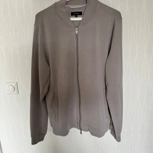 Grå skön tröja från Martinique. - Snygg grå tröja från Matinique med dragkedja framtill. Jackan har en stilren design med långa ärmar och ribbade muddar vid ärmslut och nederkant. Perfekt för en casual look.