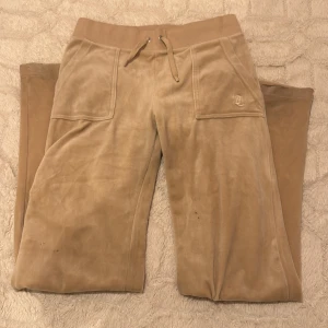 Beige mjukisbyxor från Juicy Couture - Snygga beige mjukisbyxor från Juicy Couture i mjukt material. Byxorna har en bekväm resårmidja med snörning och praktiska fickor både fram och bak. Perfekta för en avslappnad stil.