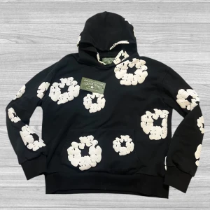 Denim Tears hoodie - Säljer en cool svart hoodie från Denim Tears med ett unikt blommönster i vitt. Hoodien har en klassisk passform med långa ärmar och en bekväm huva. Perfekt för att ge din outfit en trendig touch!