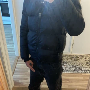 Svart dunjacka från Schott - Säljer en svart dunjacka från Schott med huva och dragkedja. Jackan har en pufferdesign och är perfekt för kyligare väder. Den har långa ärmar och två fickor med dragkedja framtill.