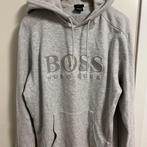Grå hoodie från Hugo Boss - Säljer en stilren grå hoodie från Hugo Boss med stor logga på bröstet. Loggan är gjort av reflex vilket synns på andra bilden vilket gör den unik! Aldrig använt denna men köpte den när jag var yngre. Storleken är barn XL, jag är 186cm och den passar perfekt. Vuxen storlek M!