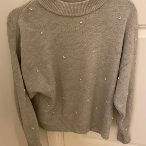 Grå tröja med pärlor från H&M - Säljer en stilren grå tröja från H&M dekorerad med små pärlor. Tröjan har en normal passform och långa ärmar, perfekt för kyligare dagar. Den är tillverkad i ett mjukt material som ger en bekväm känsla. 