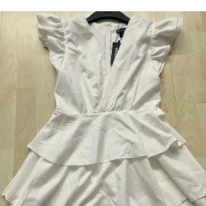 Studentklänning med volanger - Säljer en söt vit klänning med volanger och v-ringning. Klänningen har korta ärmar med puffiga detaljer och en omlott-design. Perfekt för sommarens alla tillfällen!