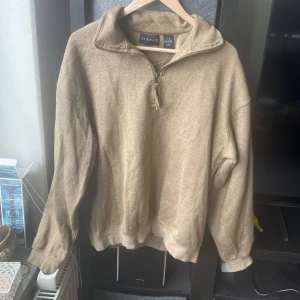 Beige tröja med dragkedja från GANT - Sönder vid ärmen!!Säljer en beige tröja från GANT i 100% bomull. Tröjan har en halv dragkedja och broderad logga på ärmen.
