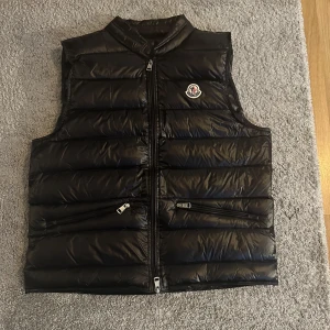 Svart dunväst från Moncler - Snygg svart dunväst från Moncler med dragkedja och två fickor framtill. Perfekt för kyligare dagar och ger en stilren look. Moncler-logga på bröstet för extra stilpoäng. Priset är inte hugget i sten kan gå ner lite med en snabb och smidig affär
