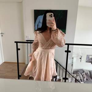 Säljer en elegant champagnefärgad klänning med glittriga paljetter på överdelen och en omlott-design. Klänningen har långa ärmar och en v-ringad hals. Kjolen är i ett lätt och flödande material. 