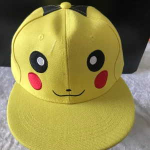 Barn Pikachu keps - Säljer en cool gul keps med Pikachu-design. Kepsen har justerbar passform och är perfekt för Pokémon-fans. Den har Pikachus ansikte med röda kinder och svarta öron på framsidan.  Helt ny 