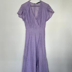 Ljuslila klänning från Zara - Säljer en vacker ljuslila klänning från Zara med kortärmad design och v-ringning. Klänningen har ett subtilt prickigt mönster och en omlott-look framtill. Perfekt för sommarens alla tillfällen!