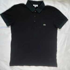 Svart pikétröja från Lacoste - Säljer en stilren svart pikétröja från Lacoste i slim fit. Tröjan har korta ärmar och en klassisk krage med tre knappar. Den ikoniska Lacoste-loggan är broderad på bröstet. Perfekt för en avslappnad men ändå elegant look.