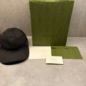 Svart keps från Gucci - Snygg svart keps från Gucci med diskret mönster och klassiska gröna och röda ränder på sidan. Kepsen har en justerbar rem baktill för perfekt passform. Perfekt för att ge din outfit en lyxig touch. Storlek m 58cm
