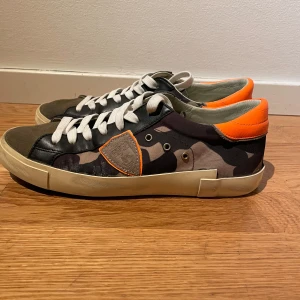 Camouflage sneakers från Philippe Model - Snygga sneakers från Philippe Model med ett coolt camouflage-mönster och orange detaljer. Skorna har en rund tå och snörning framtill. Perfekta för dig som vill sticka ut med stil.