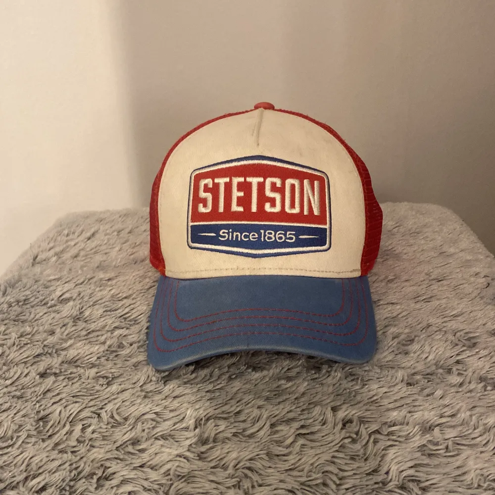 Snygg keps från Stetson med en cool retrostil. Framsidan är beige med ett stort Stetson-märke i rött och blått. Skärmen är blå och baksidan är i rött meshmaterial för bra ventilation. Perfekt för en avslappnad look! Storlek junior. Den är använd, men jag tycker att den ser väldigt fräsch ut. . Asusteet.