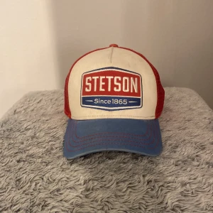 Stetson keps med röd och blå design - Snygg keps från Stetson med en cool retrostil. Framsidan är beige med ett stort Stetson-märke i rött och blått. Skärmen är blå och baksidan är i rött meshmaterial för bra ventilation. Perfekt för en avslappnad look! Storlek junior. Den är använd, men jag tycker att den ser väldigt fräsch ut. 