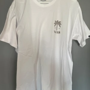 Vit t-shirt med palmtryck från Vans - Säljer en vit t-shirt från Vans med ett coolt palmtryck på bröstet och ett större tryck på ryggen. Perfekt för en avslappnad stil. T-shirten har korta ärmar och är gjord i mjuk bomull.