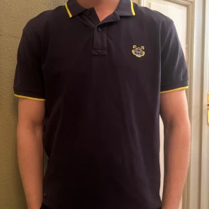 Svart pikétröja från Kenzo - Snygg svart pikétröja med gula detaljer på krage och ärmslut. Tröjan har en broderad logga på bröstet och knappar vid halsen. Perfekt för en stilren look. Märket Kenzo.