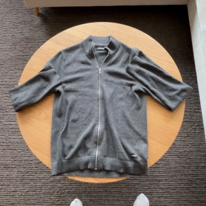 Grå kofta från Jack & Jones - Säljer en stilren grå kofta från Jack & Jones med dragkedja framtill. Koftan har en ribbad krage och trekvartsärmar, perfekt för lager-på-lager under kyligare dagar. Skicket är 9,5 / 10 utan defekter.  Priset är inte hugget i sten - skriv vid minsta fundering.