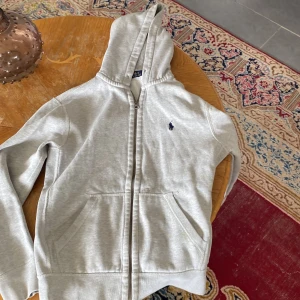 Grå hoodie från Polo Ralph Lauren - Säljer en stilren grå hoodie från Polo Ralph Lauren med dragkedja och huva. Tröjan har en broderad logotyp på bröstet och praktiska fickor framtill. Perfekt för en avslappnad look.