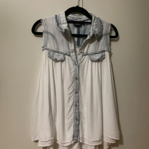 Vit ärmlös blus från Only - Säljer en vit ärmlös blus från Only med jeansdetaljer. Blusen har en krage och knappar framtill samt två bröstfickor. Perfekt för en avslappnad stil med en touch av denim.