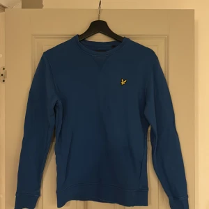 Blå tröja från Lyle & Scott - Säljer en stilren blå tröja från Lyle & Scott med deras ikoniska logga på bröstet. Tröjan har långa ärmar och en rund halsringning. Perfekt för en avslappnad look. Pris kan diskuteras!