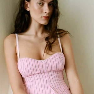 Rosa randig korsettopp - Säljer en söt rosa korsettopp med smala axelband och vertikala ränder. Toppen har en figurnära passform och är perfekt för en stilren look.