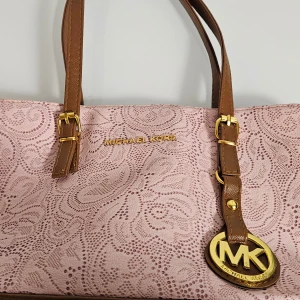 Rosa axelväska från Michael Kors - Snygg rosa axelväska från Michael Kors med ett elegant mönster och bruna detaljer. Väskan har guldfärgade spännen och en praktisk dragkedja upptill. Perfekt för att ge din outfit en lyxig touch.