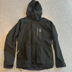 Svart vindjacka från Haglöfs - Säljer en svart vindjacka från Haglöfs med huva och dragkedja. Jackan har en bröstficka med dragkedja och två stora fickor framtill. Tillverkad i Gore-Tex-material för extra skydd mot väder och vind. 