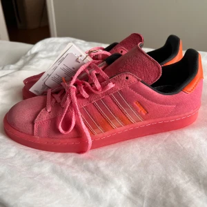 Rosa Adidas sneakers campus 44 - Snygga rosa Adidas sneakers i mocka med klassiska ränder på sidan. Skorna har snörning och en kontrasterande orange häl. Perfekta för en färgglad stil. Oanvända helt nya, finns extra snören som är svarta. Storlek 44 2/3. 