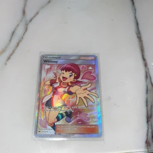 Pokémonkort - Whitney Trainer - Säljer ett Pokémonkort med karaktären Whitney, en Trainer-kort med holografisk yta. Kortet har en färgglad design med Whitney i en energisk pose. Perfekt för samlare eller Pokémon-entusiaster!