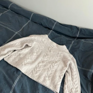 Beige stickad tröja från 157 - Säljer en mysig beige stickad tröja från 157. Tröjan har ett flätmönster och långa ärmar, perfekt för kyliga dagar. Den är både stilren och bekväm, idealisk för höst och vinter.