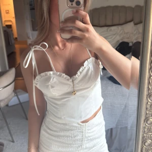 Vit croppad linnetopp med volangdetaljer - Säljer en söt vit croppad linnetopp med volangdetaljer och knytband på axlarna. Toppen har en smickrande v-ringning och smockad rygg för extra komfort. Perfekt för varma dagar och somriga outfits.