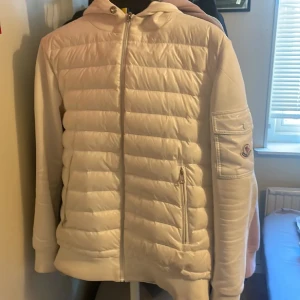  Moncler cardigan  - Säljer en stilren vit pufferjacka från Moncler med huva och dragkedja framtill. Jackan har quiltad framsida, ficka med dragkedja på ärmen och Moncler-logga på vänster arm. Perfekt för kyliga dagar.