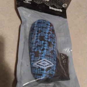 Blå och svarta benskydd från Umbro - Säljer ett par benskydd från Umbro i storlek XS. De har ett blått och svart rutmönster med Umbro-logga framtill. Benskydden har elastiska svarta sleeves för att hålla dem på plats under match eller träning. Priset kan diskuteras.