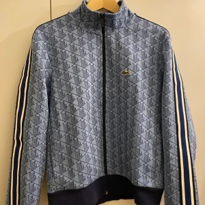 Blå mönstrad track jacket från Lacoste - Säljer en blå track jacket från Lacoste med geometriskt mönster och klassisk krokodillogga på bröstet. Jackan har dragkedja, hög krage och randiga detaljer längs ärmarna. Perfekt för en sportig och stilren look.