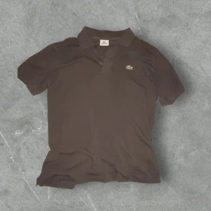 Svart pikétröja från Lacoste - Säljer en svart pikétröja från Lacoste med klassisk krage och två knappar framtill. Tröjan har korta ärmar och den ikoniska gröna krokodilloggan broderad på bröstet. Perfekt för en stilren och avslappnad look.