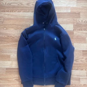 Mörkblå hoodie från Polo Ralph Lauren - Säljer en klassisk mörkblå hoodie från Polo Ralph Lauren med dragkedja och huva. Tröjan har två stora fickor framtill och den ikoniska loggan broderad på bröstet. Perfekt för en avslappnad stil.