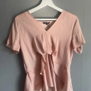 Ljusrosa blus med volang och v-ringning - Säljer en ljusrosa blus från Vero Moda med v-ringning och fina volangdetaljer framtill. Blusen har korta ärmar och en lös passform som ger en avslappnad och feminin look.