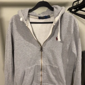 Grå hoodie med dragkedja från Polo Ralph Lauren - Grå hoodie från Polo Ralph Lauren med dragkedja och vit dragsko i huvan. Klassisk röd logga broderad på bröstet och två fickor framtill. Perfekt för en avslappnad stil.