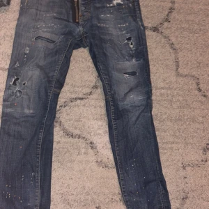 Blå slitna jeans från Dsquared2 - Säljer ett par blå jeans från Dsquared2 med slitningar, hål och färgstänk. Jeansen har dragkedja och knapp med Dsquared2-logga. Klassisk femficksmodell och smal passform. Perfekta för dig som gillar en edgy streetstil.