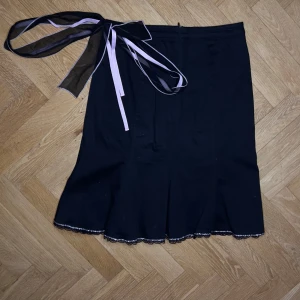 Svart kjol med rosett och spetsdetalj - Säljer en svart kjol med vacker volang och spetskant nertill. Kjolen har en stor dekorativ rosett i svart och ljusrosa band i midjan. Storlek liten S. 100% bomull 