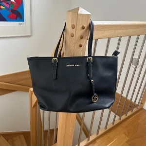 Michael Kors väska - MICHAEL KORS JET SET TRAVEL SAFFIANO TOTE! Svart Michael Kors-väska med gulddetaljer. Har inga slitningar i axelband dock lite märken på det guldiga MK-märket. Väskan har 4 innerfack och en med dragkedja. Perfekt för skolan eller jobb, får plats med dator och matlåda etc. Style # 30S4GTVT2L. 