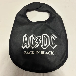 Haklapp ACDC - Svart haklapp med vit AC/DC Back in Black-tryck framtill. Tillverkad i ett slätt tygmaterial med rundad form och enkel knäppning i nacken. Perfekt för små rockfans.
