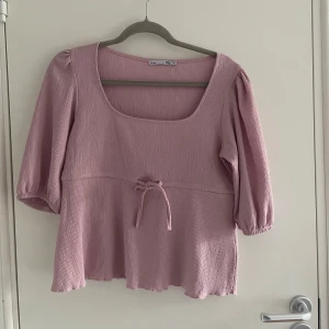 Ljusrosa blus med knyt från lager 157 - Supersöt ljusrosa blus med fyrkantig ringning och trekvartsärmar. Blusen har ett knyt i midjan och är tillverkad i ett ribbat material som ger en fin struktur. Perfekt för vår och sommar! 