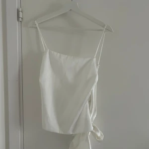 Vit satin topp med knyt från Zara - Säljer en elegant vit topp från Zara i satinliknande material. Toppen har smala axelband och knytdetalj i sidan som ger en snygg siluett. Perfekt att styla till jeans eller kjol.