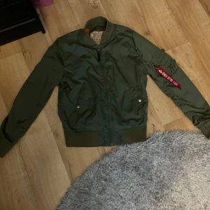 Grön bomberjacka från Alpha Industries - Säljer en klassisk grön bomberjacka från Alpha Industries med orange foder och ikonisk 'Remove Before Flight'-tag på ärmen. Jackan har dragkedja, ribbade muddar och fickor med tryckknappar. Perfekt för dig som gillar streetwear och tidlös stil. Ny pris är 1500 