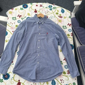 Blårutig skjorta från Ralph Lauren - Snygg blårutig skjorta från Ralph Lauren med klassisk button-down krage och röd broderad logga på bröstet. Skjortan har lång ärm och vita knappar framtill. Perfekt för dig som gillar stilrena och tidlösa plagg.