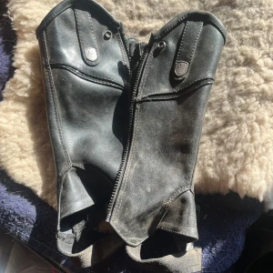 Svarta skinnchaps från Mountain Horse - Säljer ett par svarta skinnchaps från Mountain Horse med dragkedja och resår. Märkets logga syns på framsidan. Perfekta för ridning och ger bra stöd och skydd för benen till de små ryttarna. Storlek 8 och 9