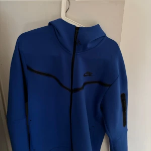 Blå hoodiejacka från Nike - Säljer en blå hoodiejacka från Nike med svarta detaljer och dragkedja framtill. Jackan har en hög krage, lång ärm med ficka på ärmen och sportig design. Perfekt för dig som gillar streetwear och sportig stil.