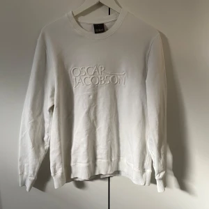 Vit sweatshirt från Oscar Jacobson - Vit sweatshirt från Oscar Jacobson med broderad logotyp på bröstet. Klassisk rund halsringning och ribbade muddar vid ärmslut och nederkant. Perfekt för en stilren och enkel look.